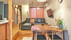 Una sala de estar acogedora con un sofá, una mesa de comedor con sillas, una lámpara colgante, un reloj de pared y un televisor montado en la pared.
