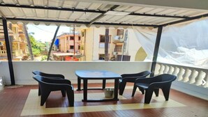 Uma área de estar coberta ao ar livre, com mesa e cadeiras, piso de azulejos e vista para edifícios ao fundo.
