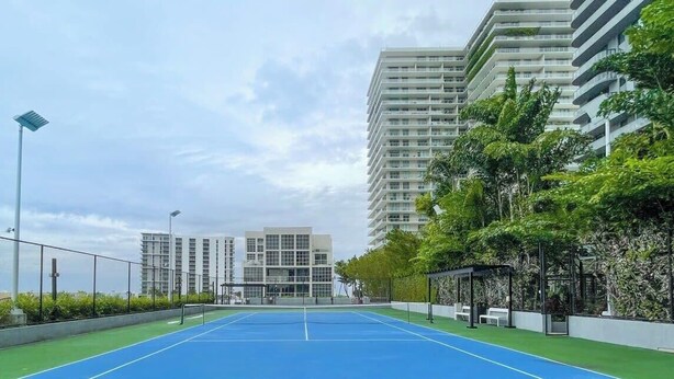 A tennis court with a green border, surrounded by a fence, and adjacent to a building with multiple balconies. - Unwind in Your Amazing City View Apartment (Miami)