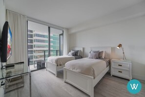 Une chambre d’hôtel moderne avec deux lits, un bureau en verre, une table de chevet avec une lampe et une vue sur la ville grâce à de grandes fenêtres.