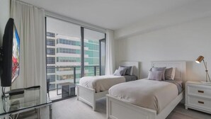 A modern hotel room with two beds, a glass desk, a nightstand with a lamp, and a view of the city through large windows. - Unwind in Your Amazing City View Apartment (Miami)