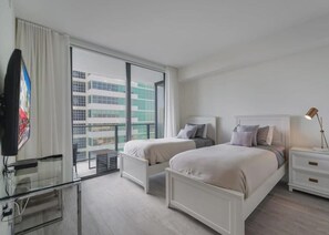 A modern hotel room with two beds, a glass desk, a nightstand with a lamp, and a view of the city through large windows. - Unwind in Your Amazing City View Apartment (Miami)