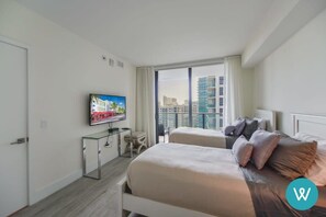 A modern hotel room with a large bed, a TV on a stand, and a view of the city through large windows. - Unwind in Your Amazing City View Apartment (Miami)