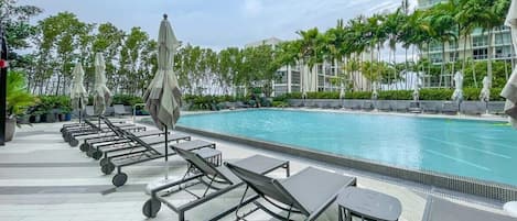 A swimming pool area with lounge chairs and umbrellas, surrounded by palm trees and a modern building.