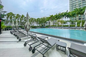 A swimming pool area with lounge chairs and umbrellas, surrounded by palm trees and a modern building. - Unwind in Your Amazing City View Apartment (Miami)