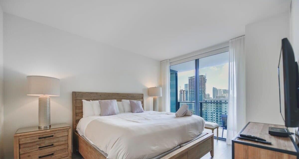 A hotel room with a large bed, bedside lamps, a desk with a computer, and a view of the city through a large window.