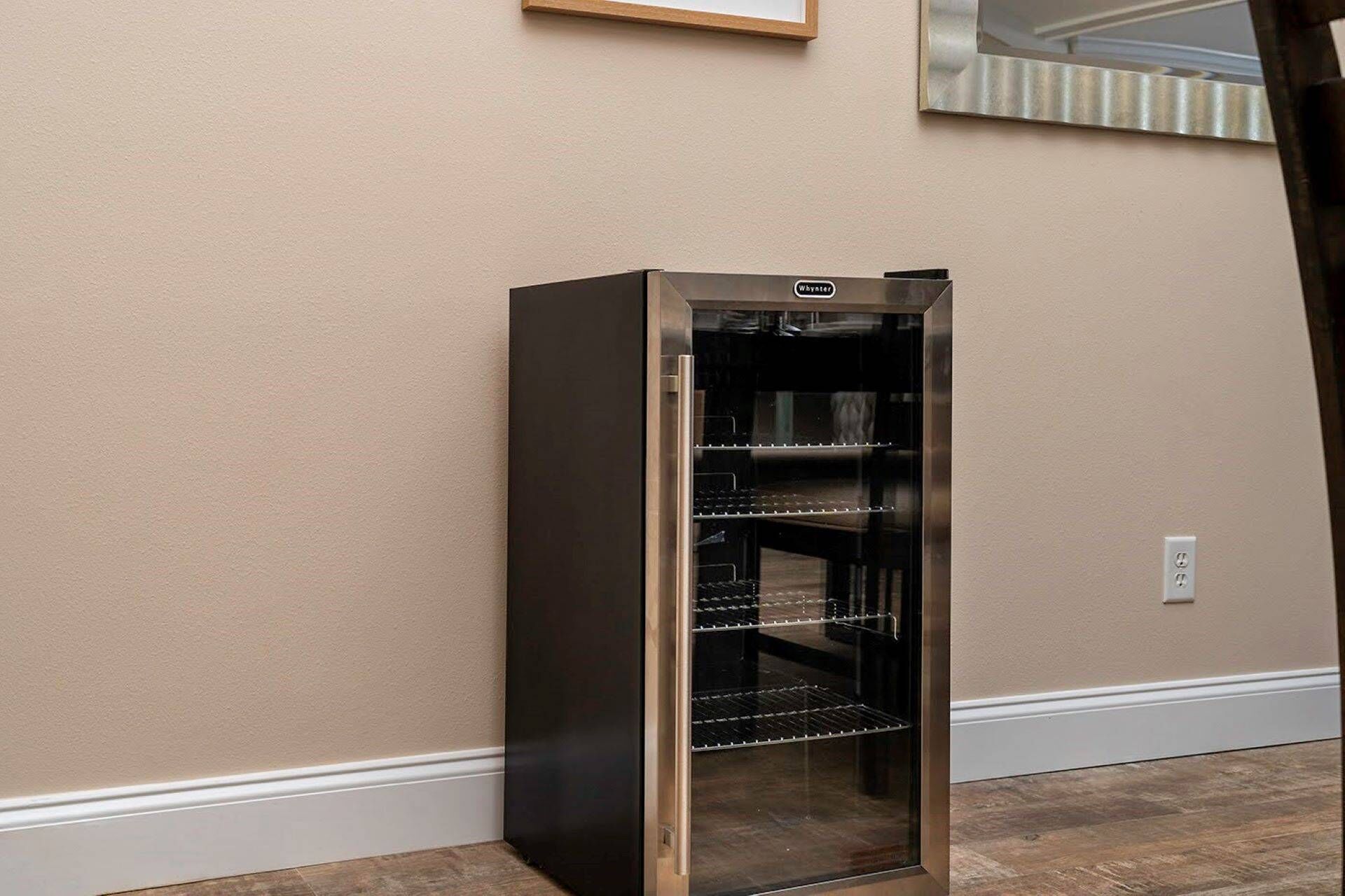 Un refrigerador de acero inoxidable con puerta de vidrio, situado en un rincón de una habitación con paredes color crema y piso de madera.