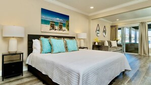 A spacious bedroom with a large bed, two bedside tables with lamps, a painting of a beach scene, and a view of the outdoors through a sliding glass door.