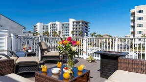 A rooftop terrace with wicker furniture, a table with drinks and a flower vase, and a view of modern buildings.
