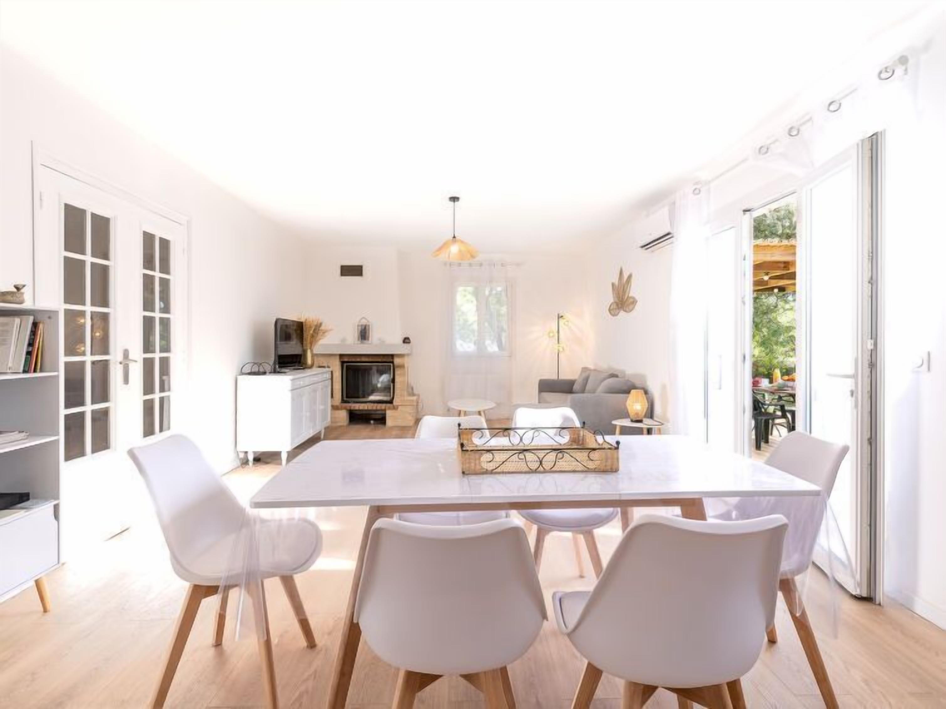 A modern, well-lit room with a dining table, white chairs, a fireplace, and a view of the outdoors.