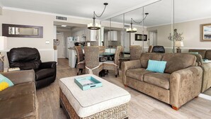 Ein Wohnzimmer mit einem braunen Ledersessel, einem braunen Sofa mit blauem Kissen, einem Couchtisch mit Tablett, Korbstühlen und einem Essbereich im Hintergrund.