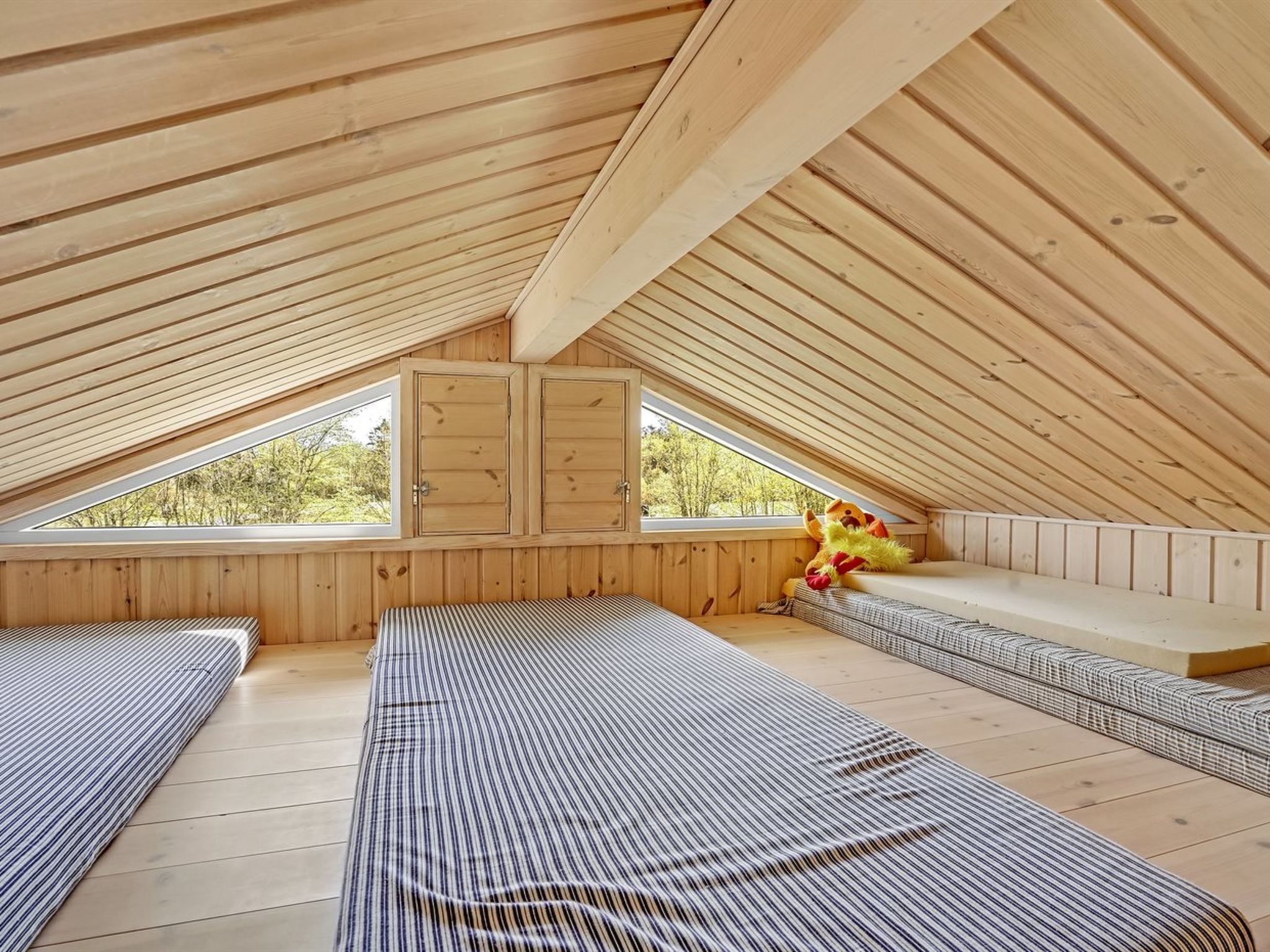 Une chambre mansardée chaleureuse avec des lambris en bois au plafond, deux lits, une fenêtre et un petit banc sur lequel est posé un jouet en peluche.