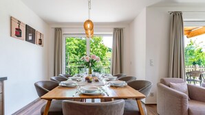 A dining area with a wooden table set for four, surrounded by chairs, with a view of greenery outside.