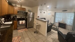 A kitchen with wooden cabinets, a stainless steel refrigerator, a microwave, and a gas stove. There is a small island with a sink and a chair. The kitchen has a tiled floor and a window with blinds. - PLACE TO BE we have everything you need  (North Port)