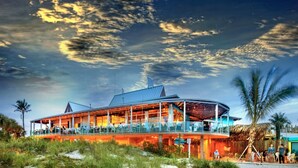 A beachfront hotel with a curved roof, outdoor seating, and a thatched roof structure nearby. - PLACE TO BE we have everything you need  (North Port)