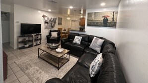 A modern living room with a black leather sofa, a glass coffee table, and a flat-screen TV. There is a dining area in the background with wooden chairs and a kitchen counter. - PLACE TO BE we have everything you need  (North Port)