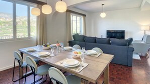 A dining area with a wooden table set for four, surrounded by chairs, and a sofa in the background. - Santa Rita House by Atlantic Holiday (Funchal)