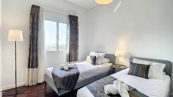 A hotel room with two beds, a window with curtains, a standing lamp, and a view of the city.