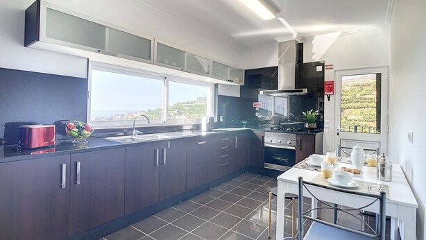 A modern kitchen with dark cabinets, a large window, and a dining area with a table and chairs. - Santa Rita House by Atlantic Holiday (Funchal)