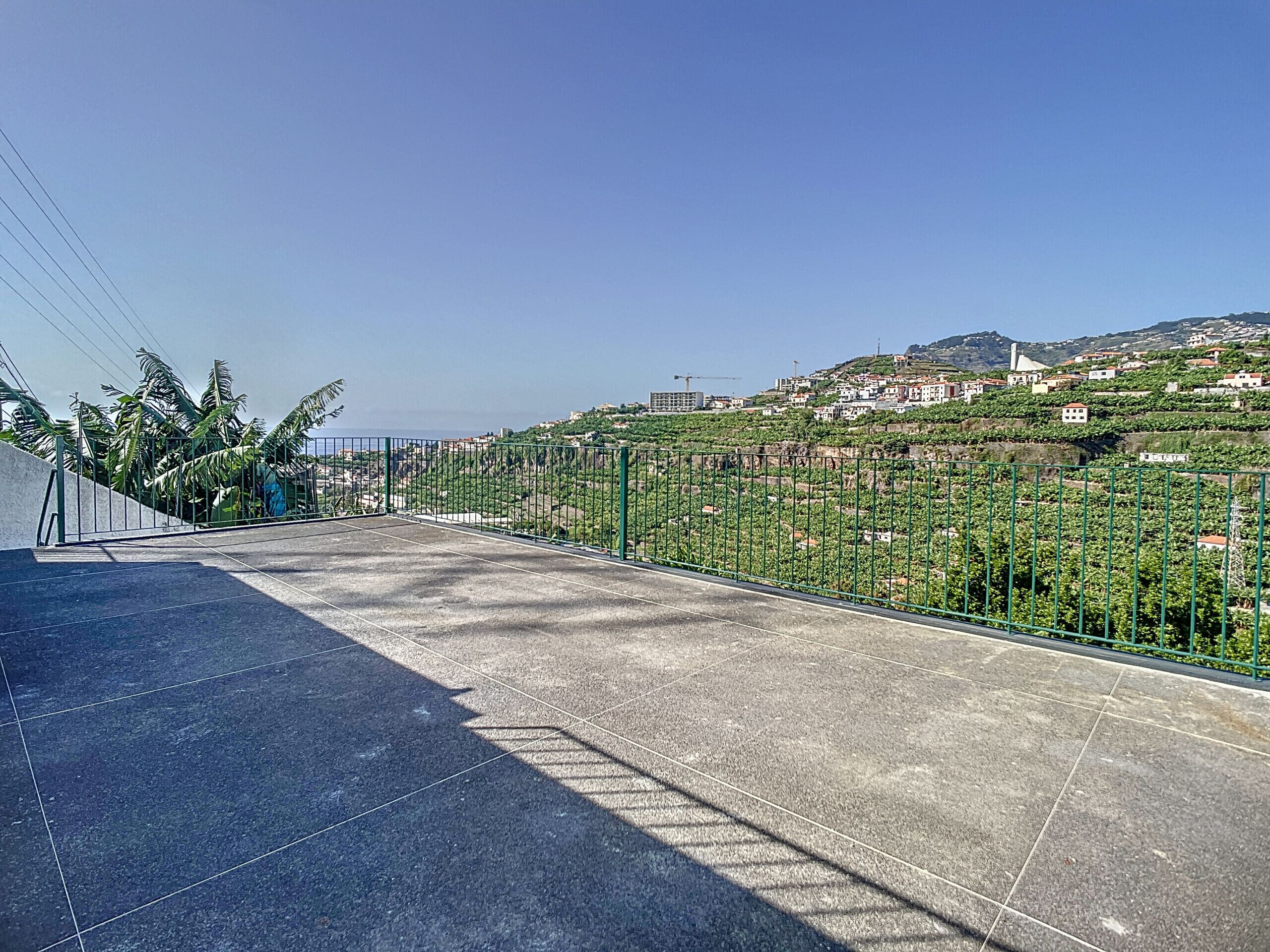 Une terrasse dallée, agrémentée d’une clôture en métal, qui offre une vue sur une colline parsemée de bâtiments et de verdure.
