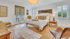 Ein Schlafzimmer mit Bett, Nachttischen, einem Sessel, einem Schreibtisch und einem großen Fenster mit Jalousien.