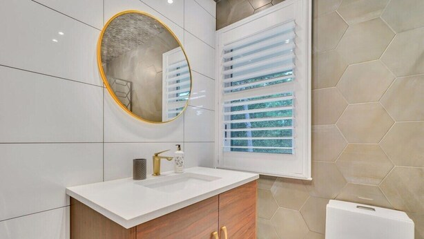 A modern bathroom with a white vanity, a round mirror, and a window with blinds. - TheGrey -Spacious,Eclectic,Home in a Serene Setting (Statesboro)