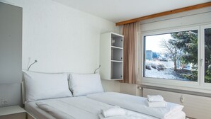 Ein ordentlich bezogenes Bett mit weißen Leinen und Handtüchern, ein Nachttisch, ein eingebautes Regal und ein Fenster mit Blick auf Bäume und ein Gebäude.