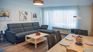 Ein modernes Wohnzimmer mit einem grauen Sofa, einem Holztisch und einem Essbereich mit Tisch für vier Personen.