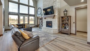 A spacious living room with a large window, a stone fireplace, a leather sofa, and a flat-screen TV mounted on the wall. - Spacious McCall Home w/ Private Lake Access! (McCall)