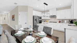 Une cuisine moderne avec des meubles blancs, des appareils en acier inoxydable et un coin repas doté d’une table en bois foncé, mise pour quatre personnes.