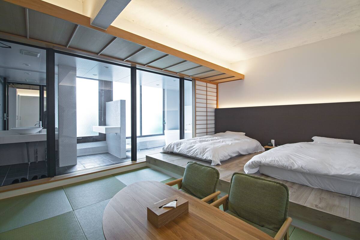 A modern hotel room with a large sliding glass door leading to a balcony, a wooden dining table, two single beds, and a small sitting area with chairs.