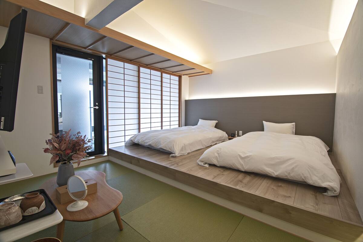 A modern hotel room with a low platform bed, a wooden desk, a small table with a vase of flowers, and a sliding shoji screen door.