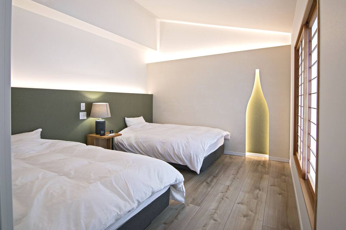 A hotel room with two beds, a bedside table with a lamp, and a large vase on the windowsill.