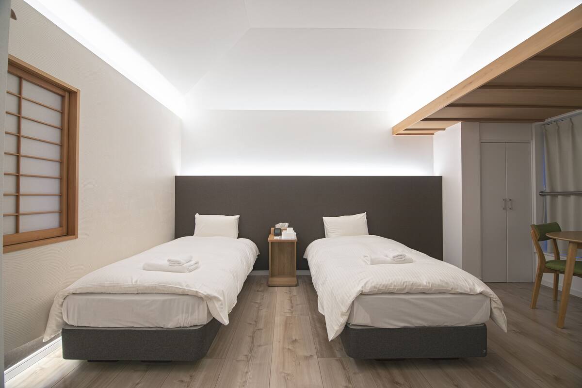 A modern bedroom with two beds, a small wooden nightstand, a green chair, and a wardrobe.