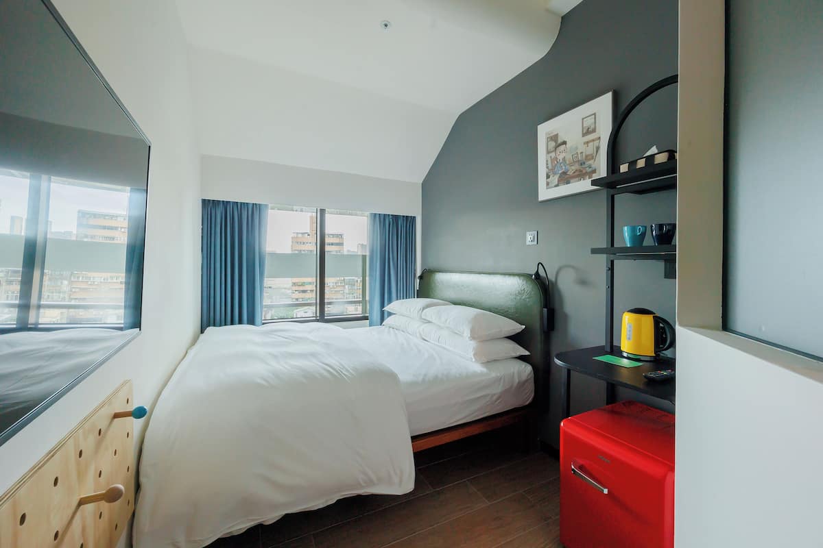 Habitación de hotel con cama, una maleta roja, estante con tazas y ventana con vista a edificios.