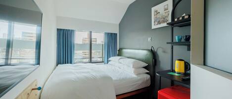 A hotel room with a bed, a red suitcase, a shelf with cups, and a window with a view of buildings.