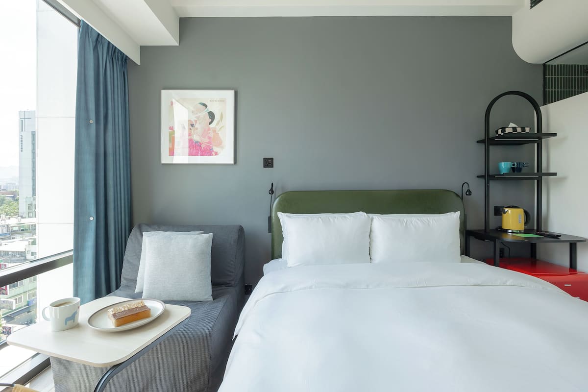 Habitación de hotel con cama, una mesita con un plato de comida y una taza, un sofá, un cuadro en la pared y una estantería con objetos.