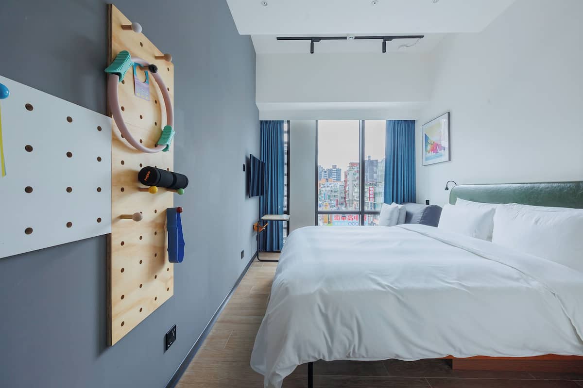 Una habitación de hotel moderna con una cama grande, un panel de ejercicios en la pared, vistas a la ciudad y un ventanal con cortinas.