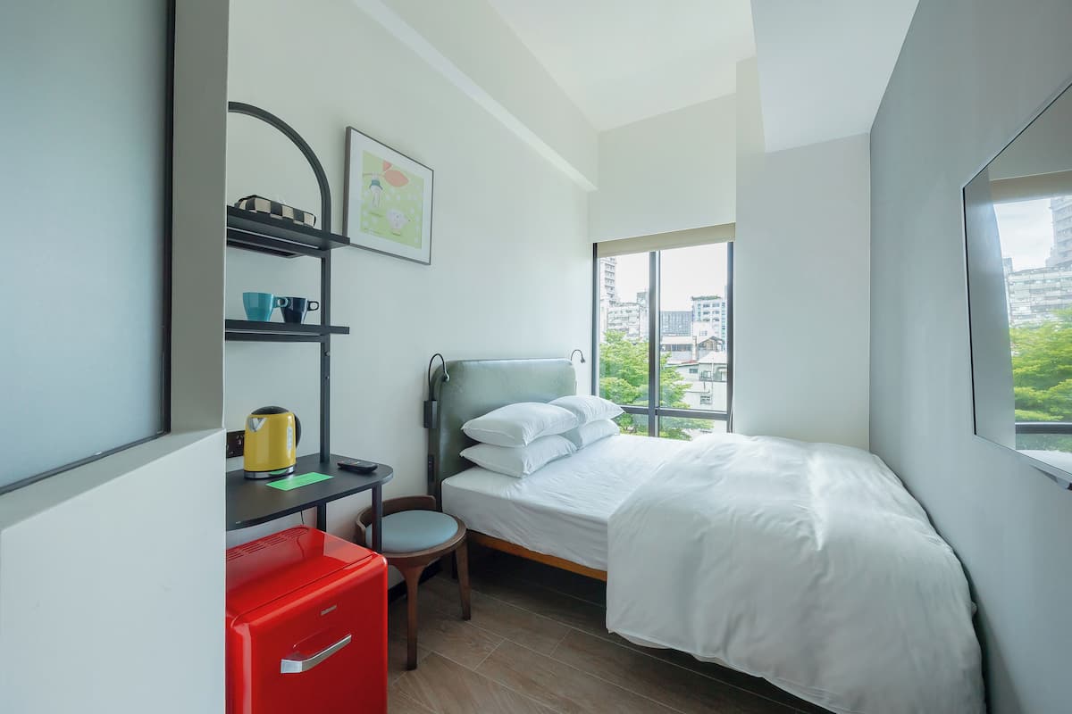 Una habitación de hotel moderna con cama, un escritorio con archivador rojo, una silla y una ventana con vistas a la ciudad.