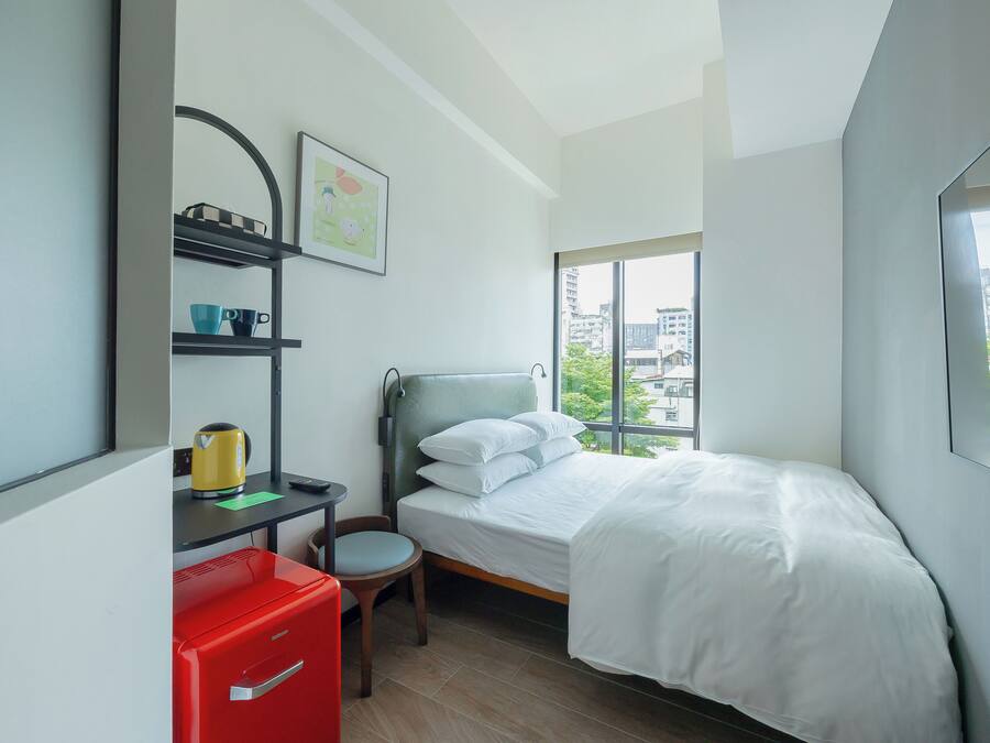 A modern hotel room with a bed, a desk with a red filing cabinet, a chair, and a window offering a city view.