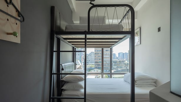 Une chambre avec un lit superposé, une vue sur la ville, un plancher en bois et une étagère fixée au mur.