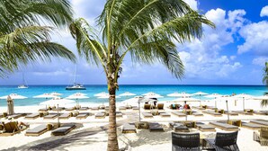Una playa con sillas de playa y sombrillas, palmeras y barcos al fondo.