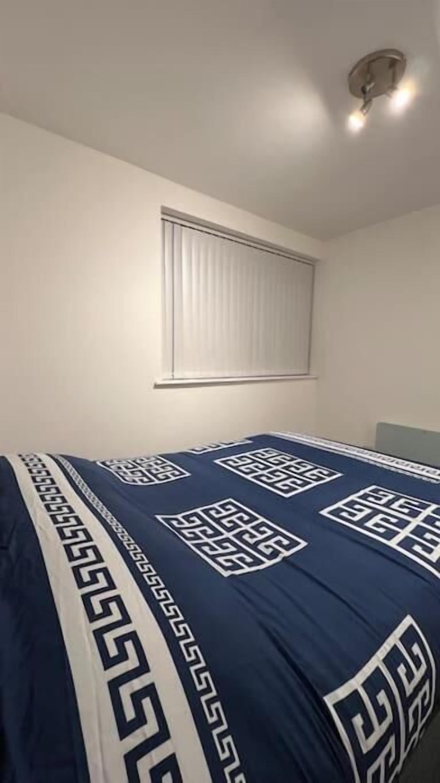 Ein Schlafzimmer mit einem Bett, das mit einer blau-weißen Bettdecke bezogen ist, einem Fenster mit Jalousien und einer Deckenleuchte.