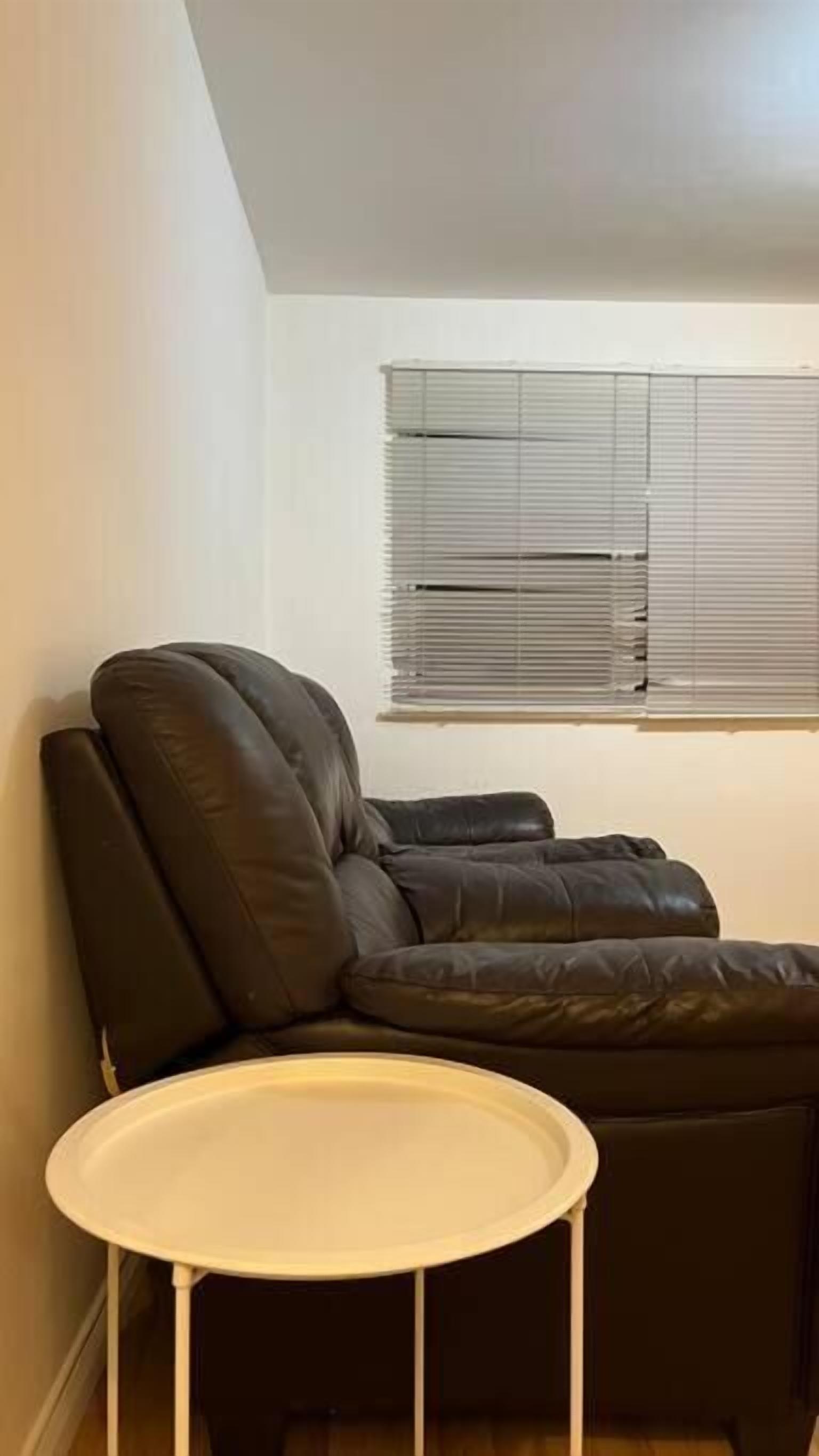 A room with a brown leather sofa, a small white table, and a window with blinds.