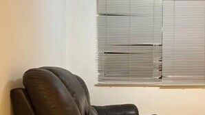 A room with a brown leather sofa, a small white table, and a window with blinds. - Charming 2bedroom Retreat in Great Barr/hamsted (Birmingham)