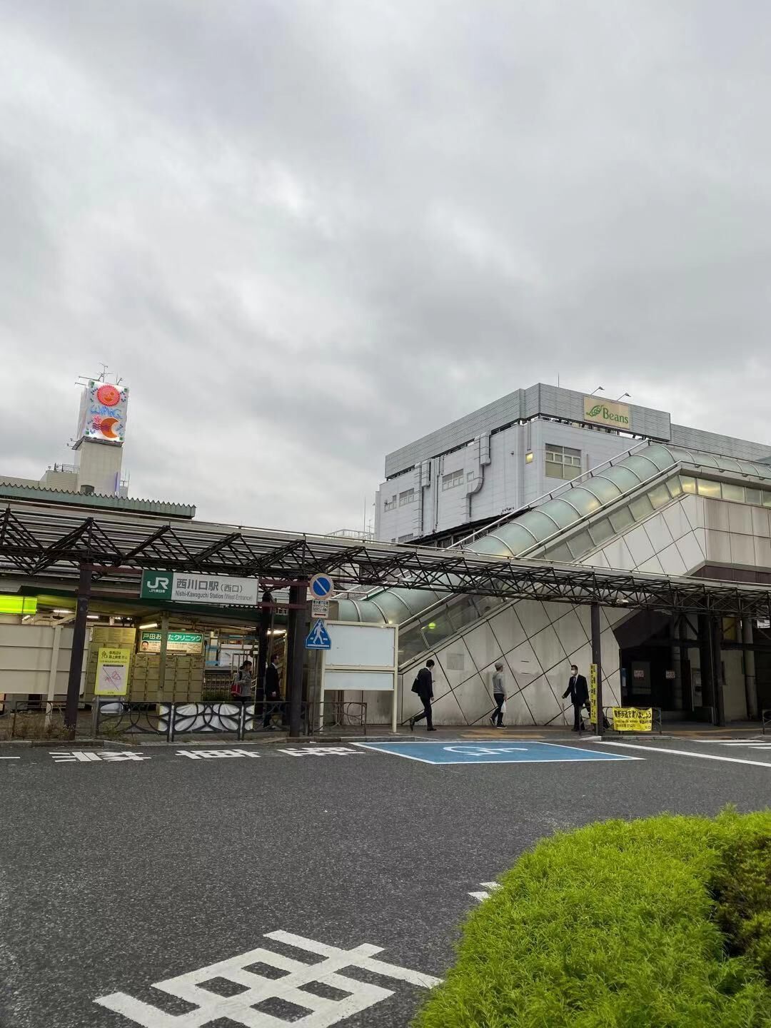 Een treinstation inkom met een modern gebouw, een voetgangersoversteek en een bord met 'Nishi-Kawaguchi Station (West Entrance)'.
