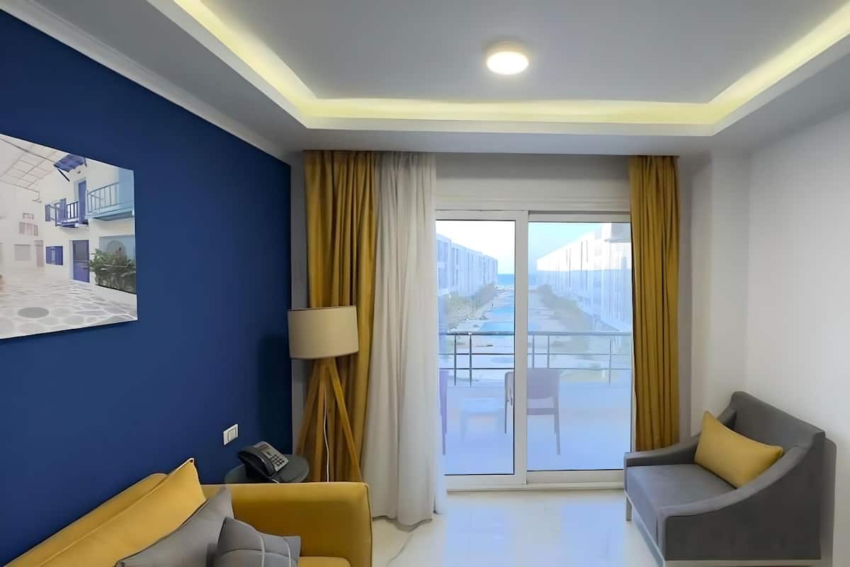 A modern hotel room with a blue accent wall, a sofa, a chair, a lamp, and a large sliding glass door leading to an outdoor balcony.
