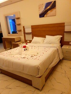 Una cama bien hecha con sábanas blancas y pétalos de rosa, un cabecero de madera, una mesita de noche y un espejo en la pared.