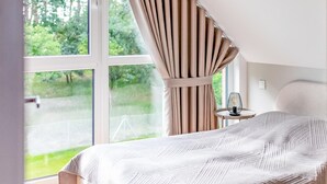 A bedroom with a large window, a bed with a patterned bedspread, a bedside table with a lamp, and curtains.