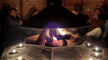 En rund ildsted i sten med ild i midten, omgivet af stearinlys, i et træpaneleret rum med højt til loftet.
